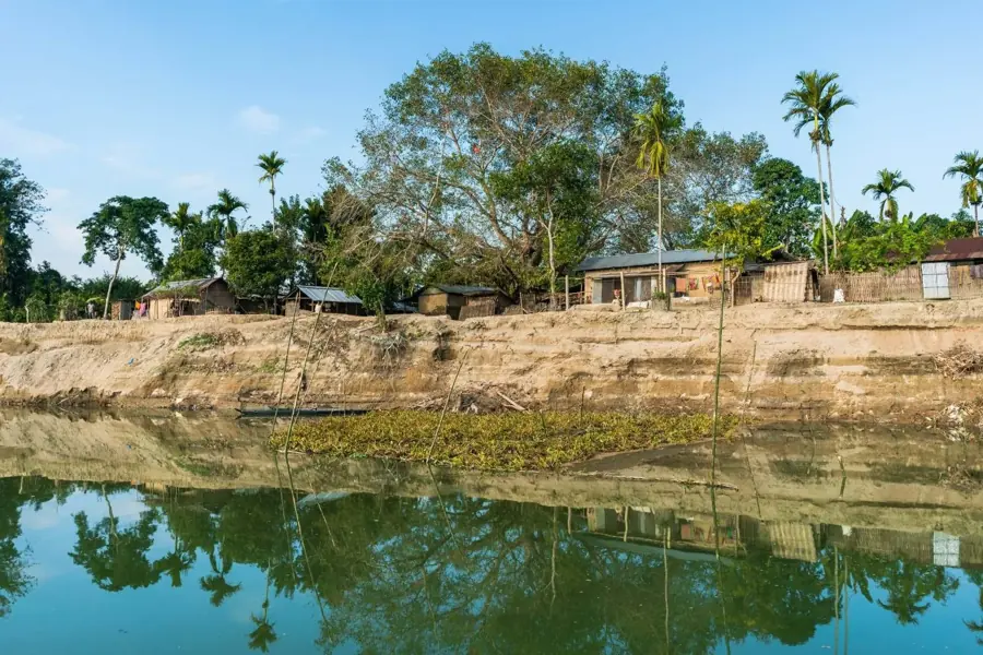 Un villaggio di case basse su una riva di terra, riflesso nell'acqua ferma di un braccio di fiume in Assam