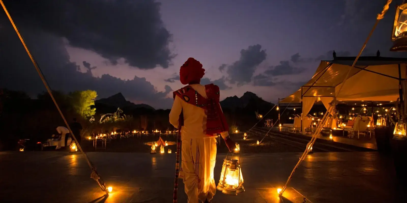 Un uomo con il turbante rosso e una lanterna, di spalle, cammina verso le tende illuminate del SUJÁN Jawai al crepuscolo