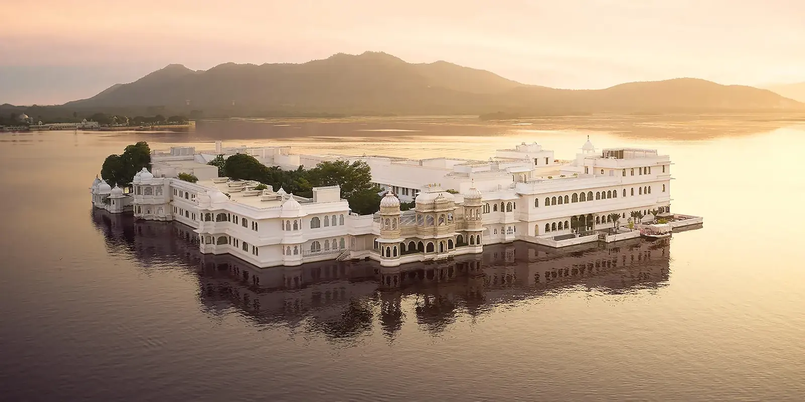 Il Taj Lake Palace, bianco, sembra galleggiare sul lago Pichola all'alba, con le colline di Udaipur all'orizzonte