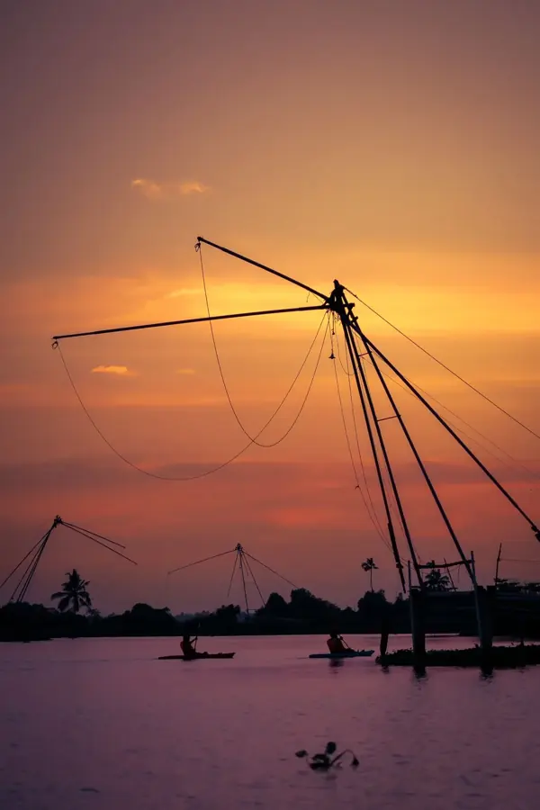 Rete da pesca cinese stagliata contro il cielo arancione del tramonto, con altre reti e piccole canoe sullo sfondo
