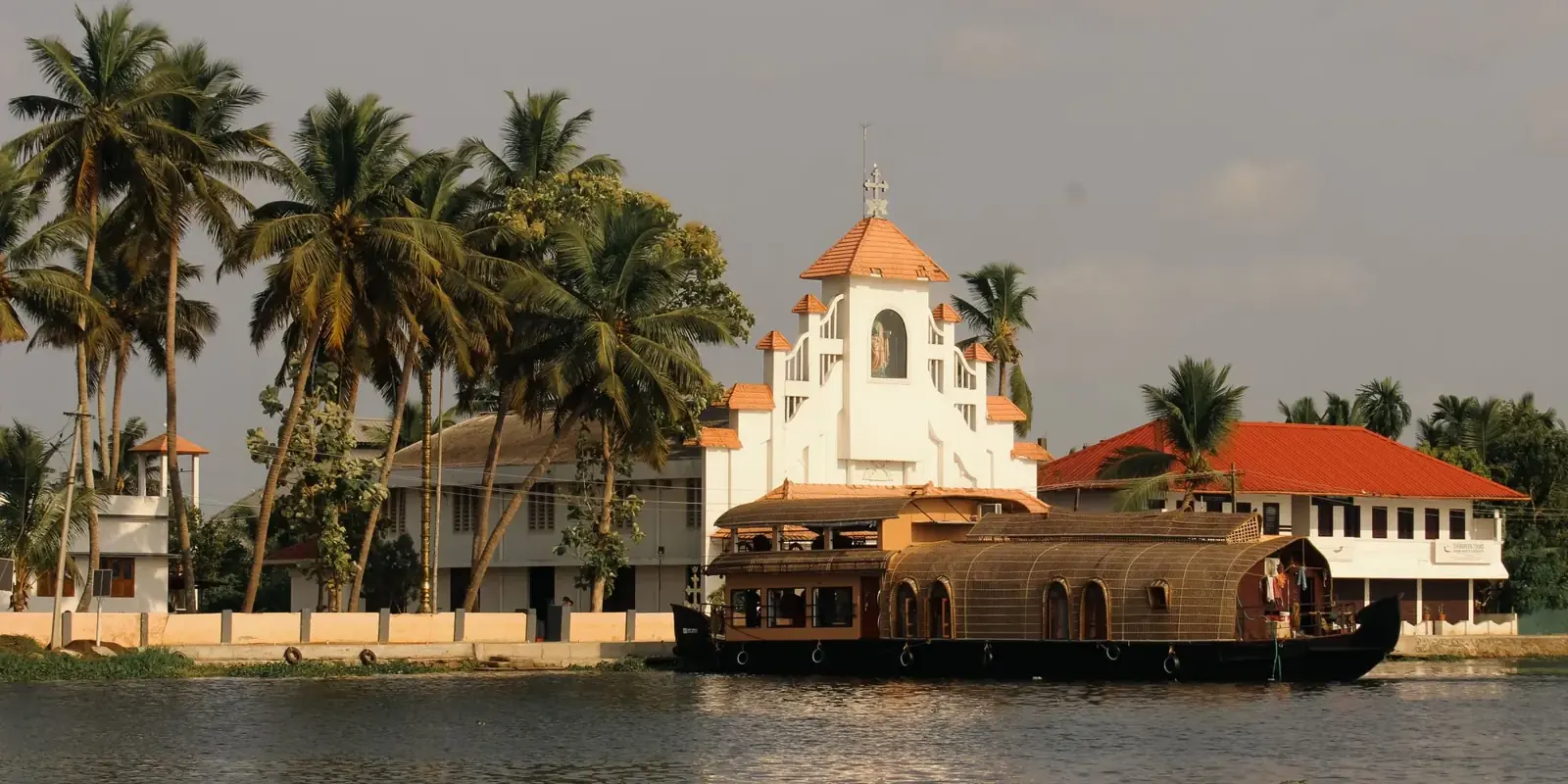Chiesa bianca tra le palme sulla riva di un grande specchio d'acqua del Kerala, con una houseboat che passa