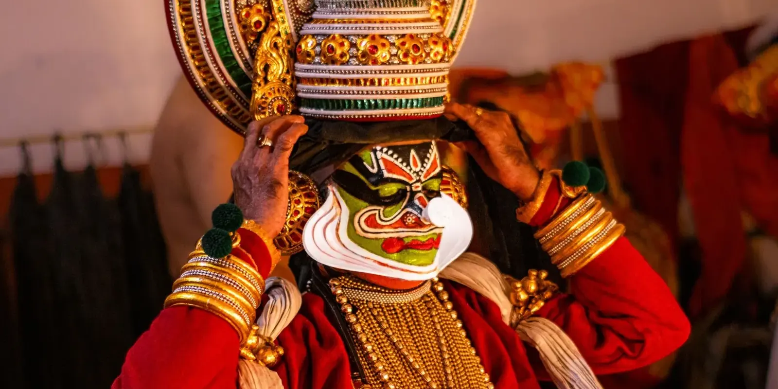 Attore di Kathakali con il volto dipinto di verde che si sistema il grande copricapo dorato dietro le quinte