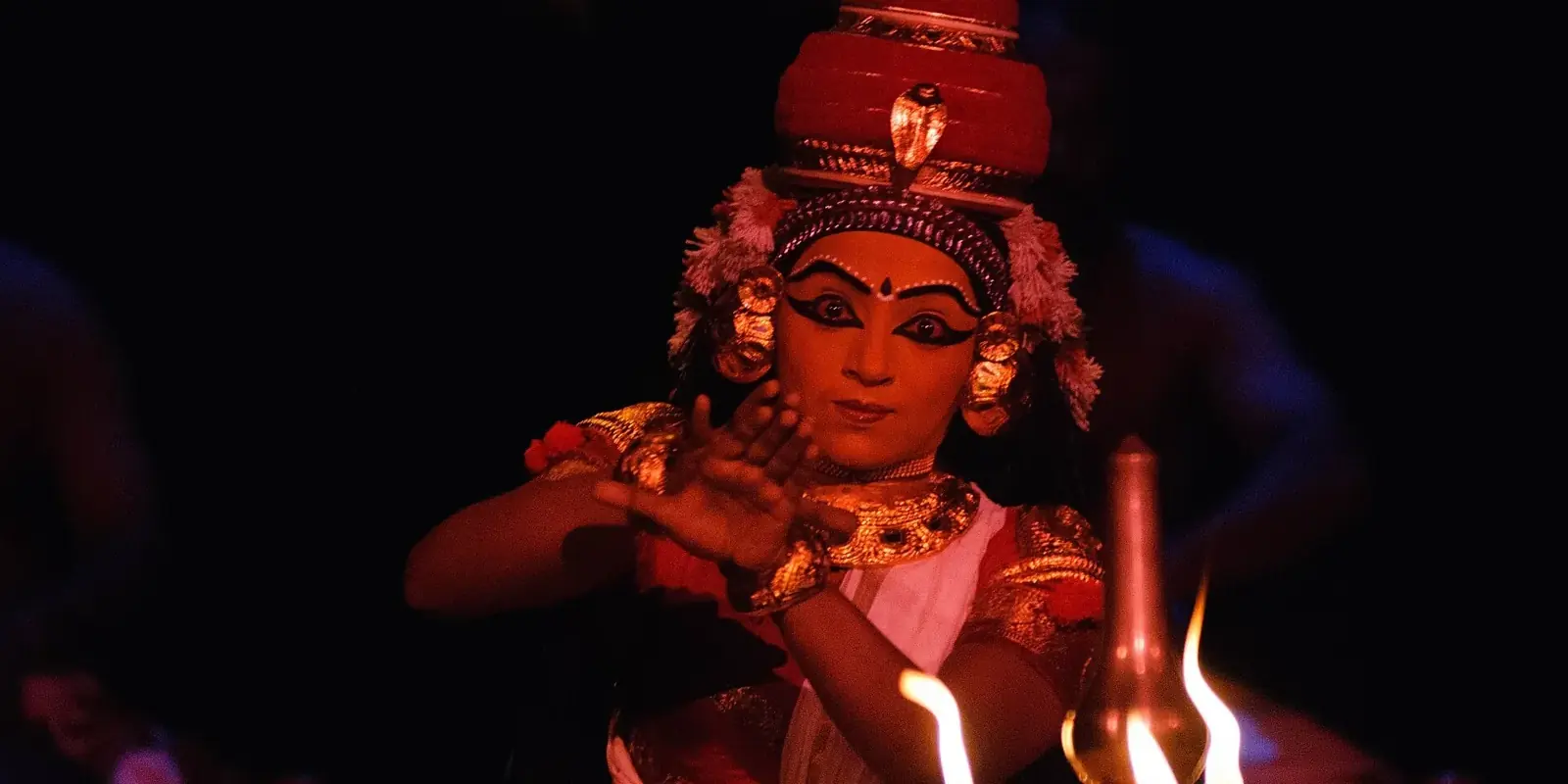 Interprete di Koodiyattam truccata e ingioiellata che danza con le mani davanti a una lampada di ottone accesa, su fondo scuro