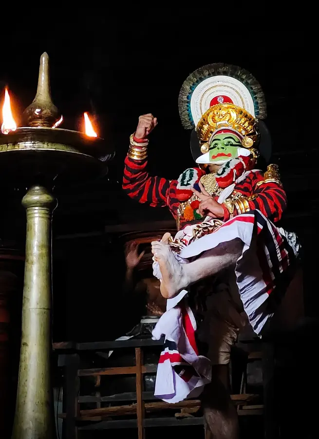 Attore di Koodiyattam con trucco verde e costume rosso e bianco accanto all'alta lampada di ottone del teatro
