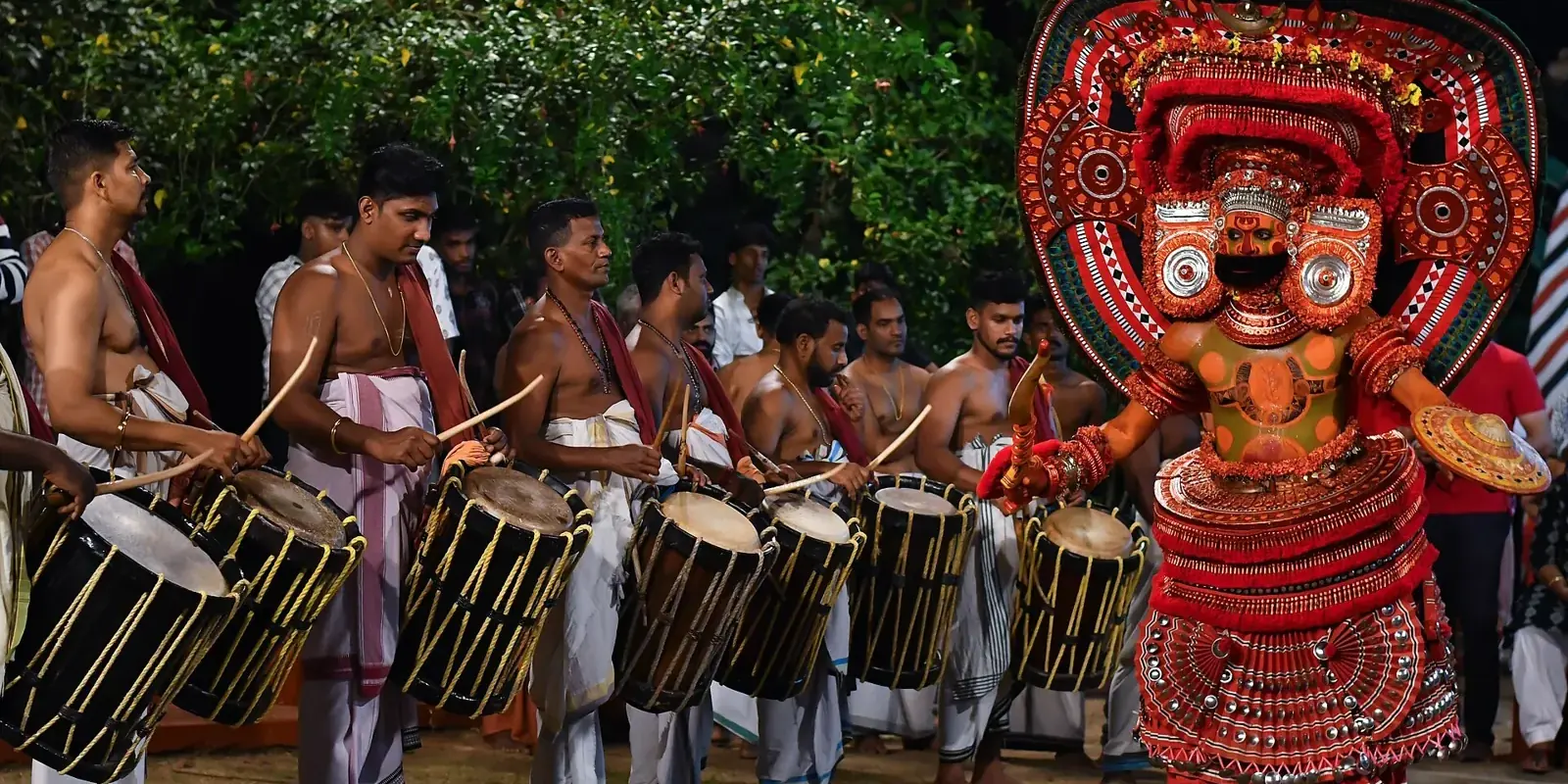 Theyyam in costume rosso con grande copricapo che danza di sera davanti a una fila di suonatori di chenda