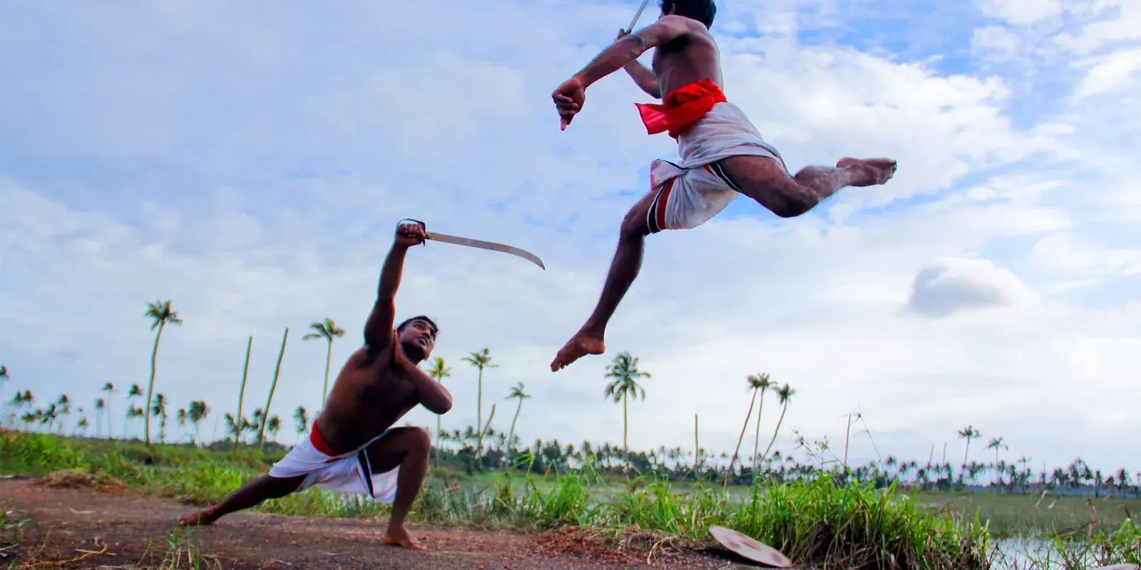 Due praticanti di Kalaripayattu con le spade, uno in salto, in un campo tra le palme del Kerala