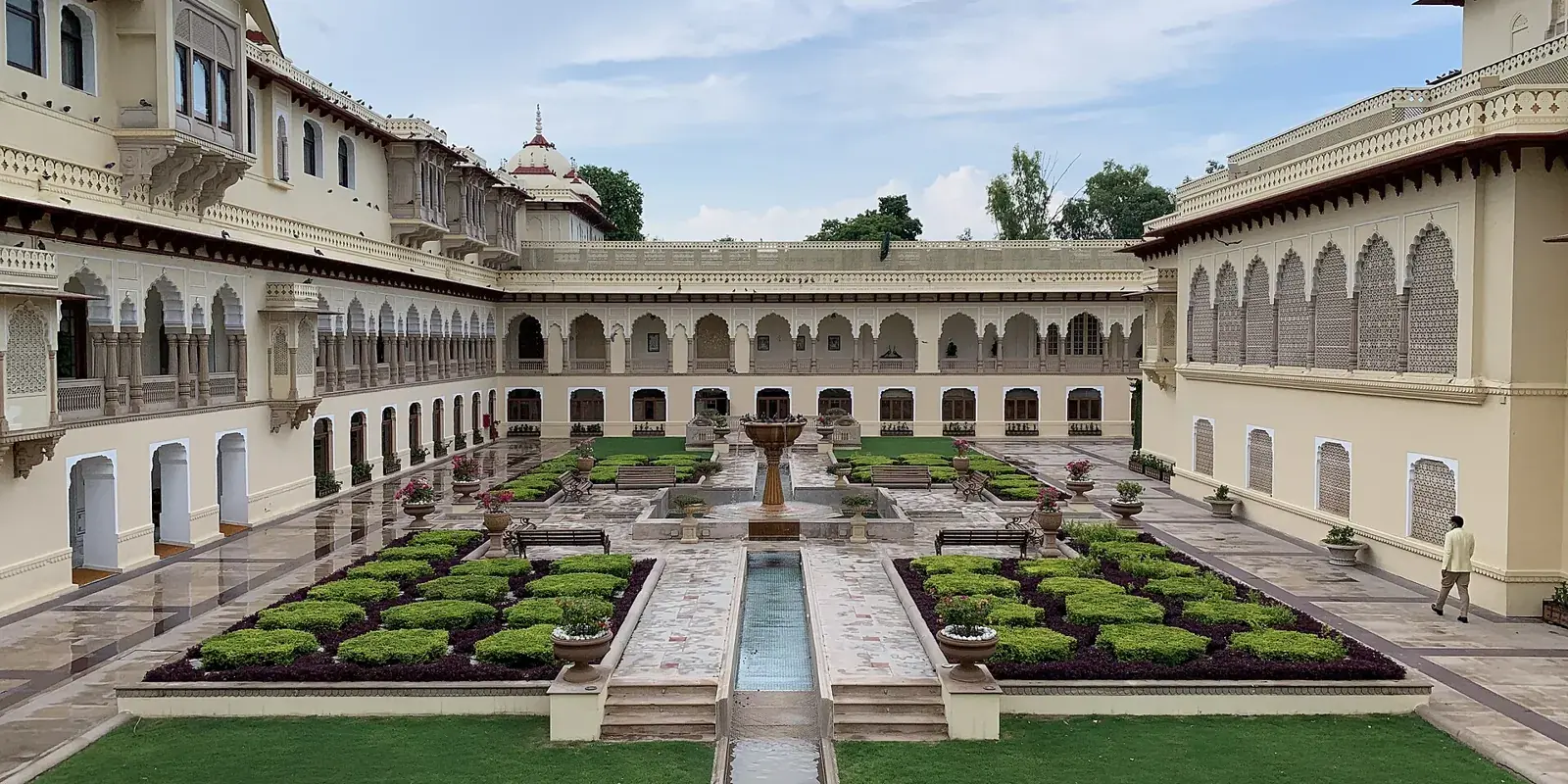 Il cortile del Rambagh Palace a Jaipur: facciate color avorio ad archi, giardino all'italiana con aiuole e una lunga vasca d'acqua