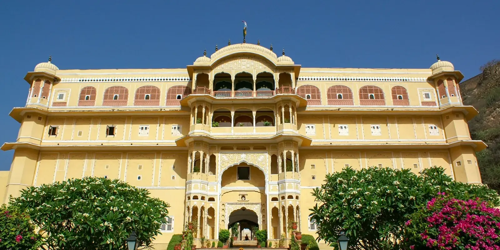 La facciata gialla del Samode Palace con la scalinata d'ingresso tra alberi in fiore, sotto un cielo blu