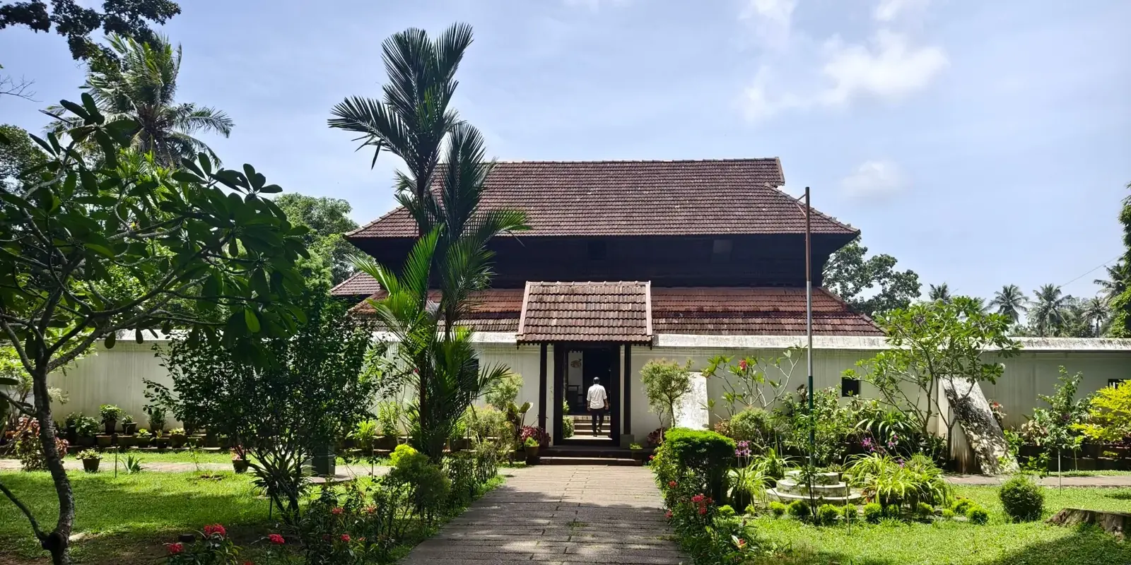 Una dimora tradizionale del Kerala con il grande tetto spiovente di tegole, in fondo a un vialetto tra prato e palme