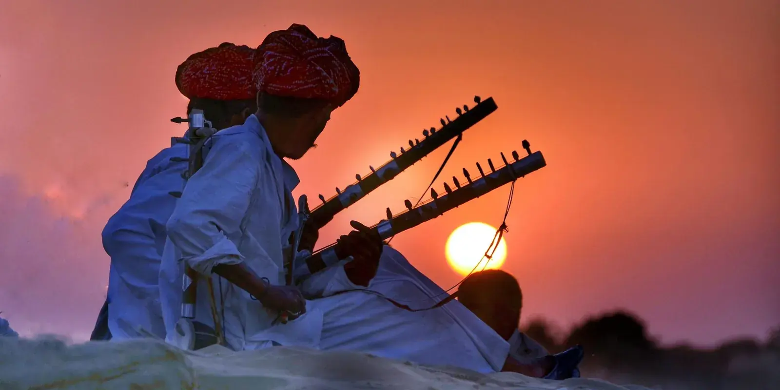 Musicisti del Rajasthan con il turbante rosso che suonano strumenti ad arco sulla sabbia, in controluce al tramonto